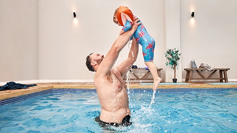 Image shows a man in a swimming pool wearing SenSura Mio Black ostomy bag. He is lifting his son up in the air. 