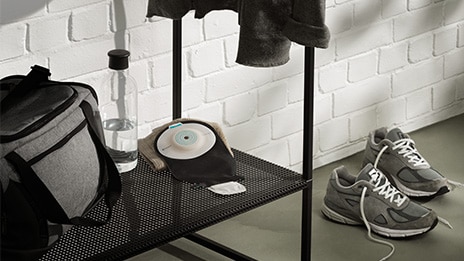 The image shows SenSura Mio black ostomy bag in a gym changing room with trainers and a water bottle